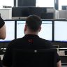 Foto von drei SAC Mitarbeitern. Ein Mitarbeiter sitzt vor drei Bildschirmen an seinem Arbeitsplatz und zwei Mitarbeiter stehen hinter ihm und Beraten sich. Alle tragen ein schwarzes SAC Polohemd