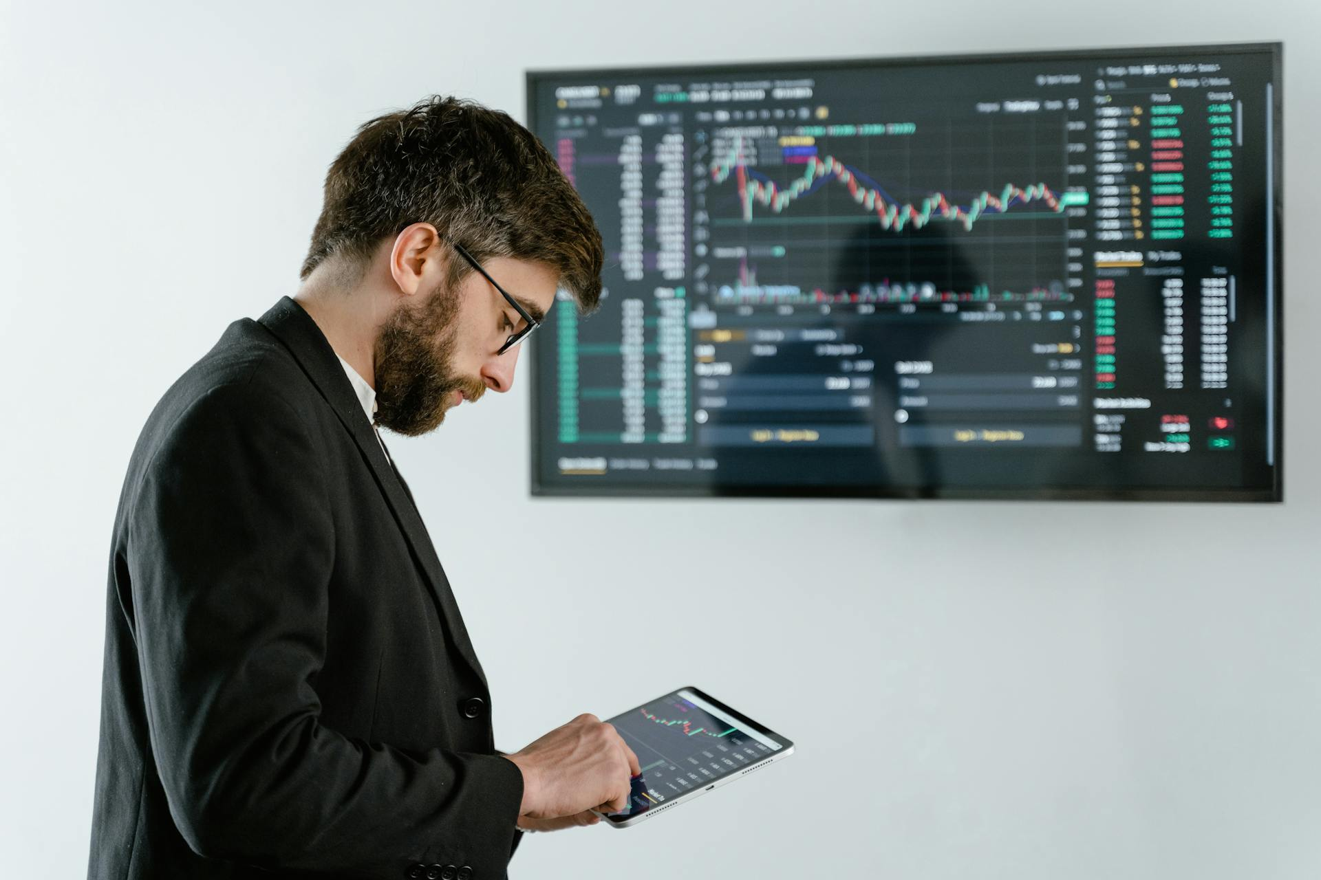 sac_beitragsbild_digital-signage-software Ein Mann in einem Anzug steht vor einer Wand mit einem großen Bildschirm. Auf dem Bildschirm sind Diagramme und Daten zu sehen, die typischerweise mit Börsenkursen oder Finanzmärkten in Verbindung stehen. Der Mann hält ein Tablet in den Händen. Das Bild steht im Zusammenhang der Digital Signage Software